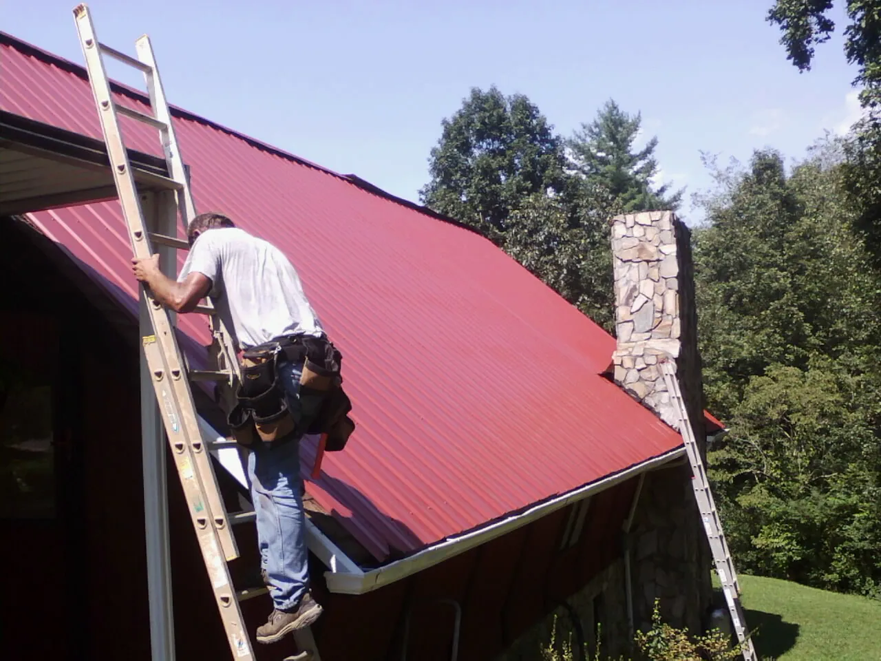 Roofer on a ladder beside a red metal roof and stone chimney — Soffit & Fascia Repair by Roofing Brothers in Stoystown